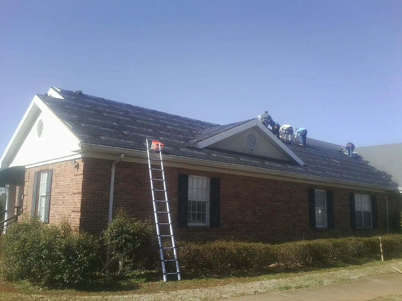 Crew tearing shingles off a brick ranch roof with a ladder set — Emergency Roof Repair work by our crew in Roslyn
